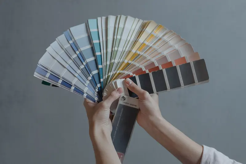 Hands holding a fan deck of colour samples in front of a grey wall.